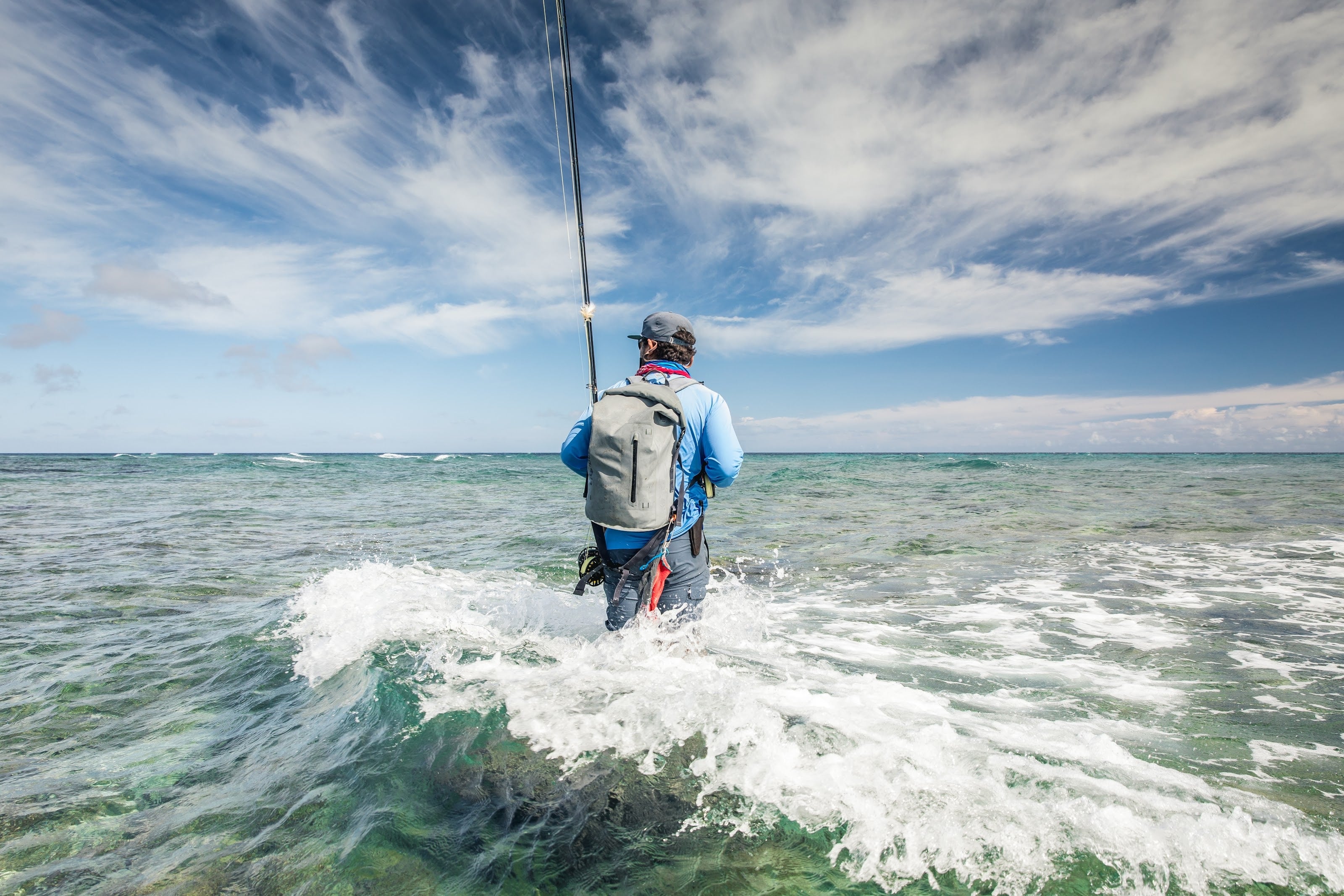 An angler with a fly rod and dry pack wading into the surf in the Seychelles