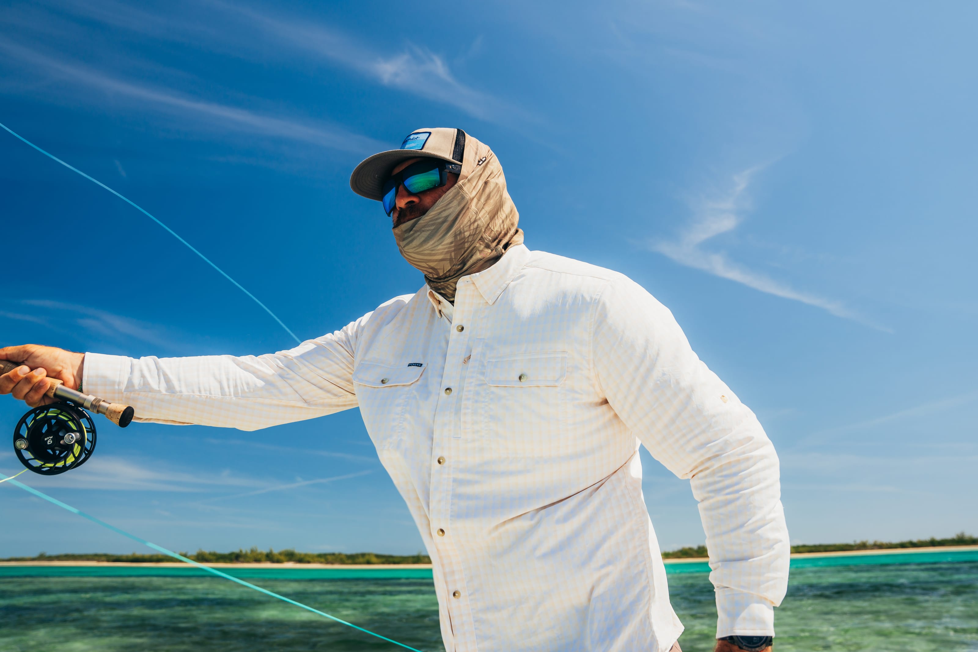 An angler in a white Poncho shirt and sun mask casting a fly rod over a turquoise saltwater flat