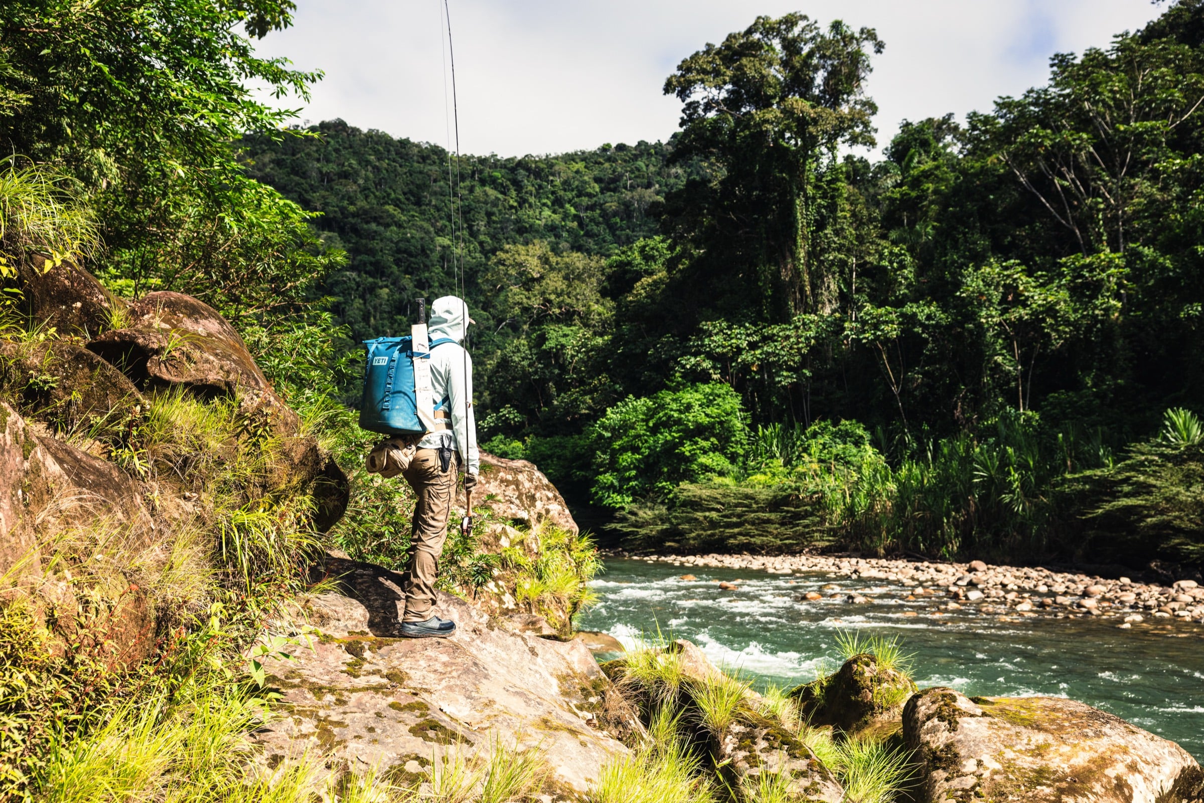 An angler scouting a jungle river with a full pack and rod